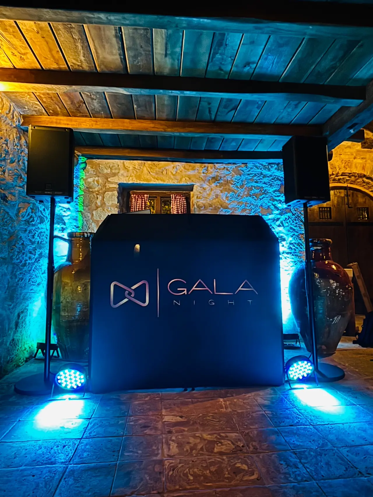 Profesionalna DJ oprema s kabinom s oznakom 'Gala Night', okružena s dva PA zvučnika na stalcima i plavim osvjetljenjem uz rustikalni kameni zid Konobe Okit - Karmelski Dvori Vodice.
